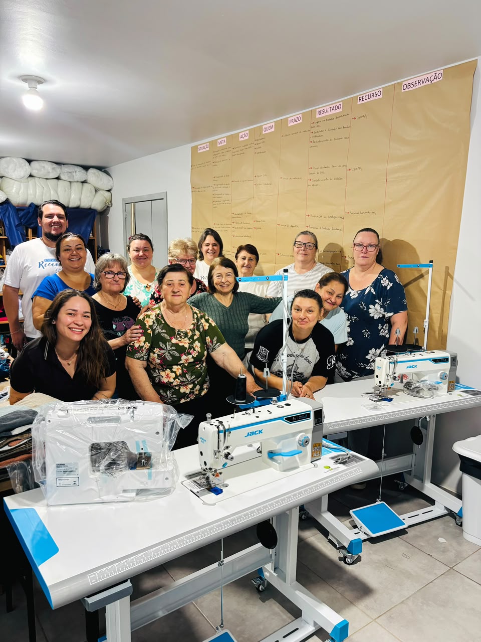 Grupo de mulheres posa junto a máquinas de costura adquiridas pelo CRAS em oficina de costura.