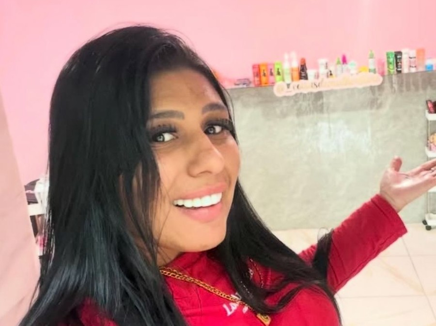Mulher sorri para câmera em ambiente interno com prateleiras de produtos ao fundo e parede rosa.