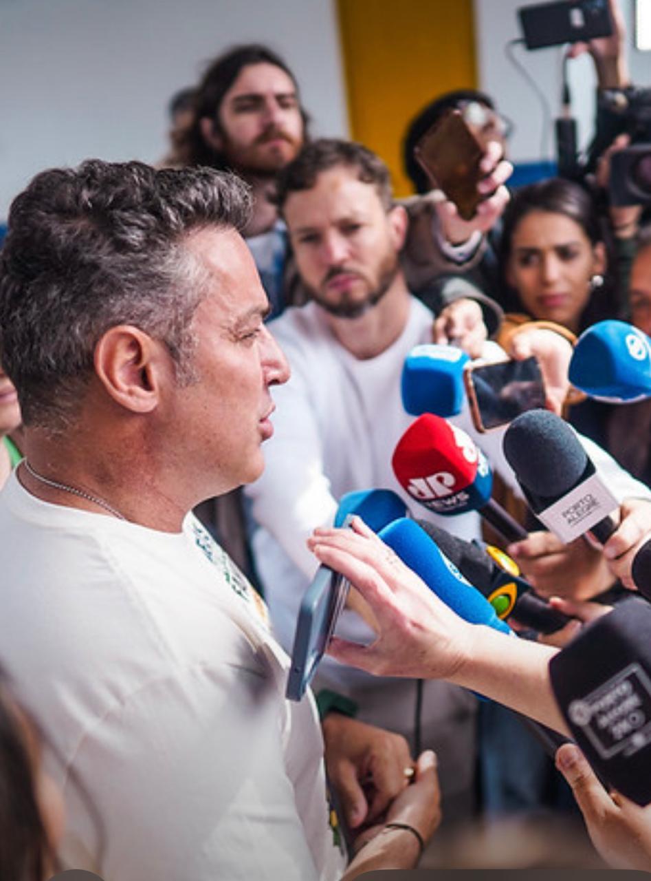 Homem em camiseta branca fala para repórteres com microfones durante coletiva de imprensa.
