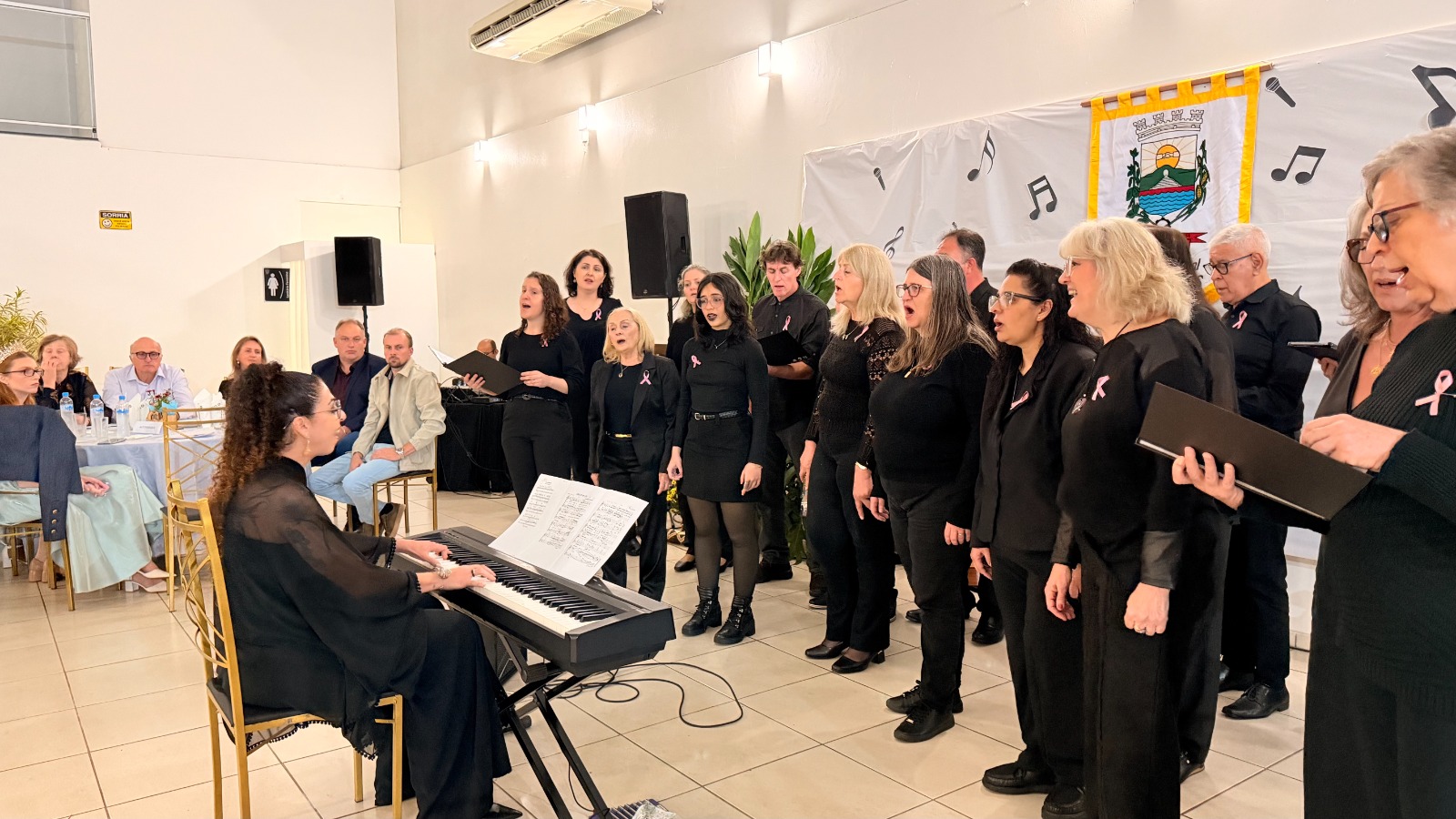 Coralistas apresentam números musicais durante o Encontro de Coros em Roca Sales, com pianista acompanhando as apresentações.
