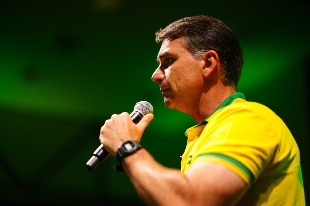 Homem em camiseta amarela e verde fala ao microfone durante evento com fundo verde.