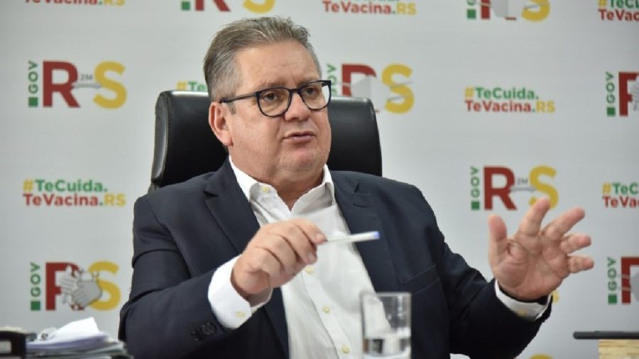 Homem de óculos e terno fala durante coletiva de imprensa com painel de fundo com logos do Rio Grande do Sul.