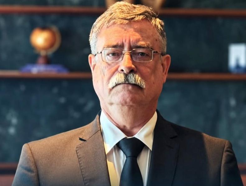 Homem de terno e gravata, com óculos e bigode, em ambiente de escritório com estantes ao fundo.