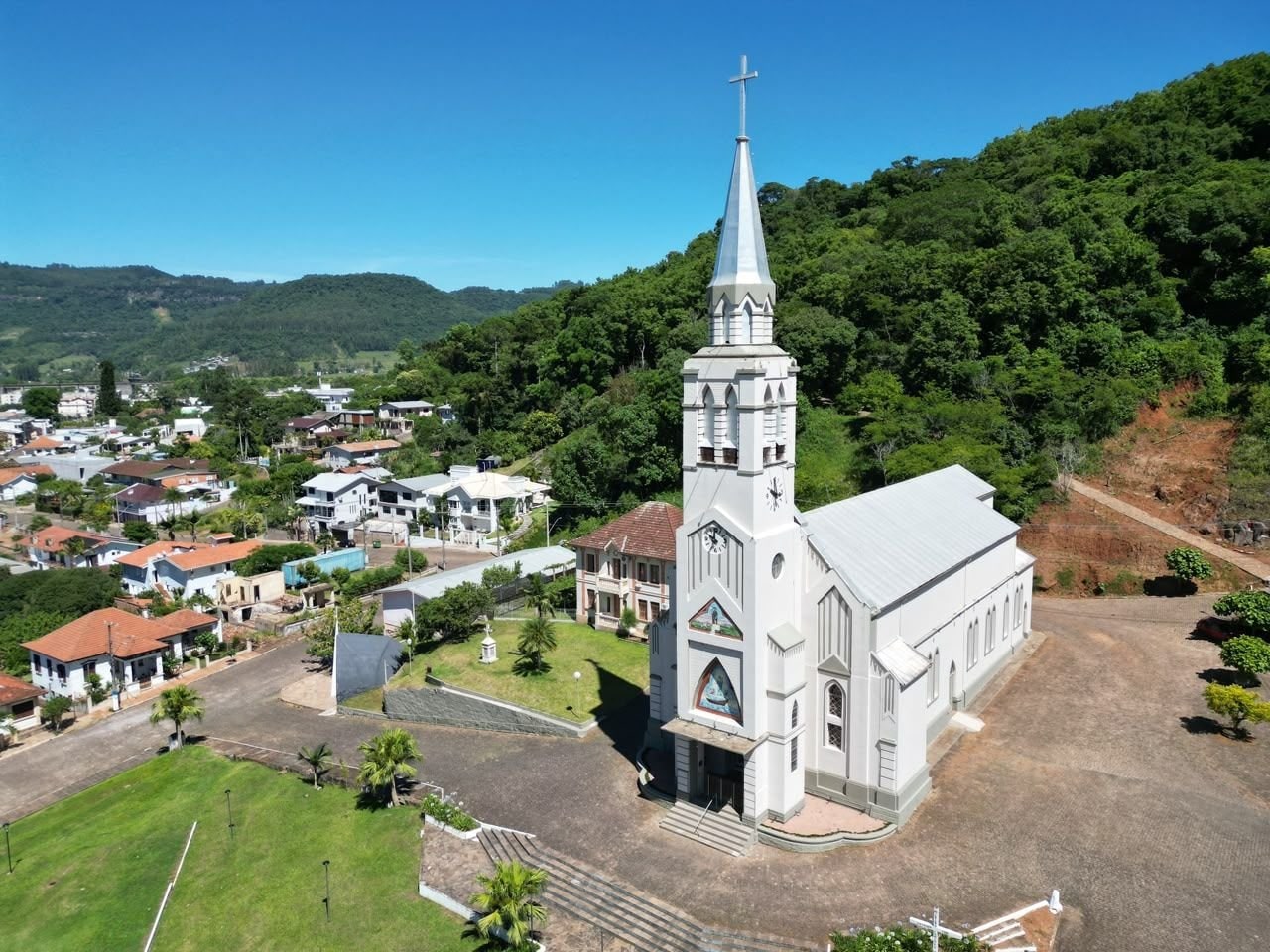 Igreja branca com torre pontiaguda se destaca na paisagem de Muçum, cercada por casarões e vegetação das encostas da região.