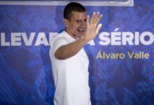 Homem em camiseta branca gesticula durante evento em estúdio com painel azul ao fundo.