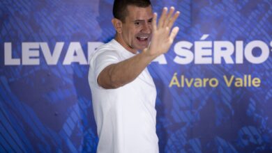 Homem em camiseta branca gesticula durante evento em estúdio com painel azul ao fundo.