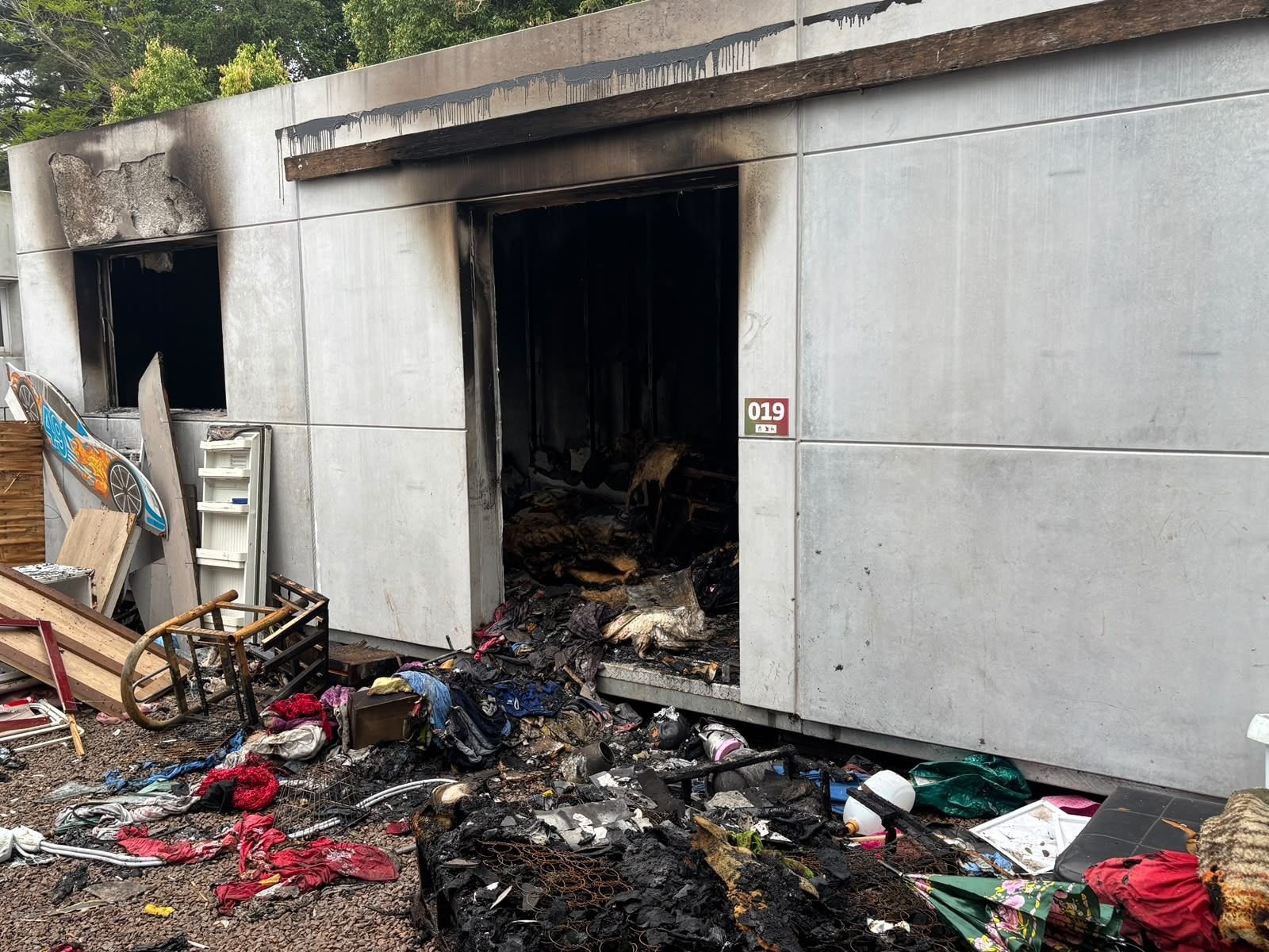 Contêiner residencial apresenta sinais de incêndio, com entulhos e roupas queimadas espalhados na entrada da moradia em Estrela.