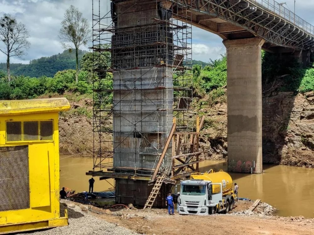 Operários trabalham na reconstrução de pilar da ponte entre Encantado e Muçum, com caminhão-betoneira abastecendo a obra às margens do rio.