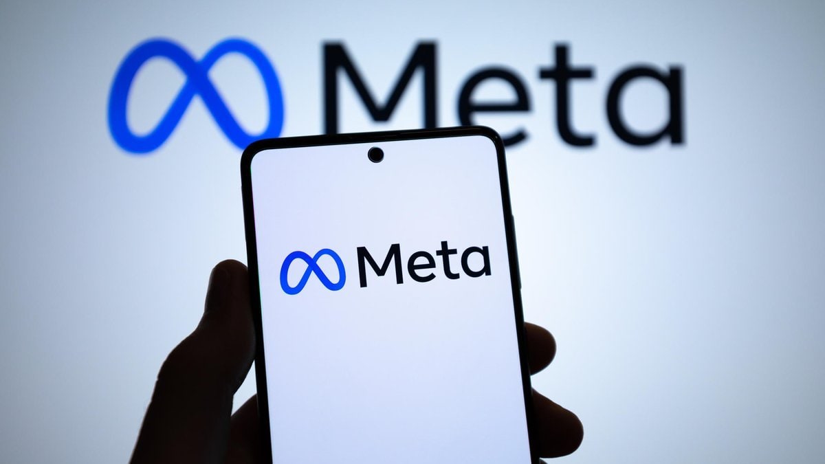 Mão segura smartphone com logo da Meta exibido na tela, com marca da empresa ao fundo.